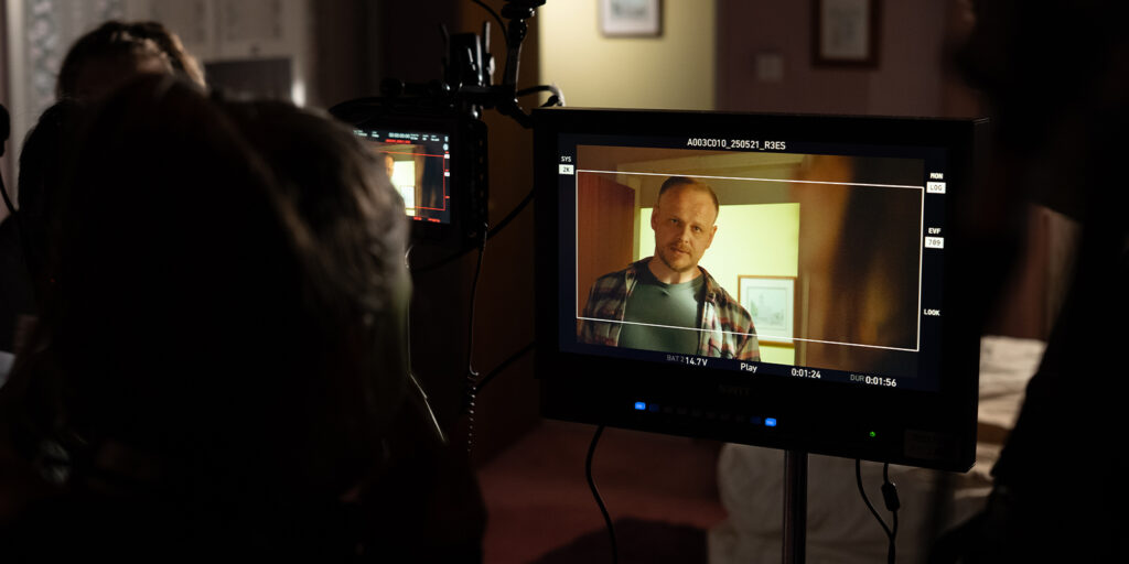 A camera monitor displays a man standing indoors, facing the camera. He wears a checked shirt over a t-shirt. Careful lighting on the film set creates balance, with people and equipment partially visible in the foreground.