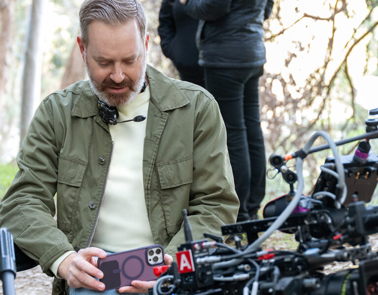 A man outdoors sits next to professional film equipment, holding a mobile phone and looking down at it. Trees and greenery are visible in the background, with another person standing behind him on the set of "Loot," filmed by Jason Oldak.