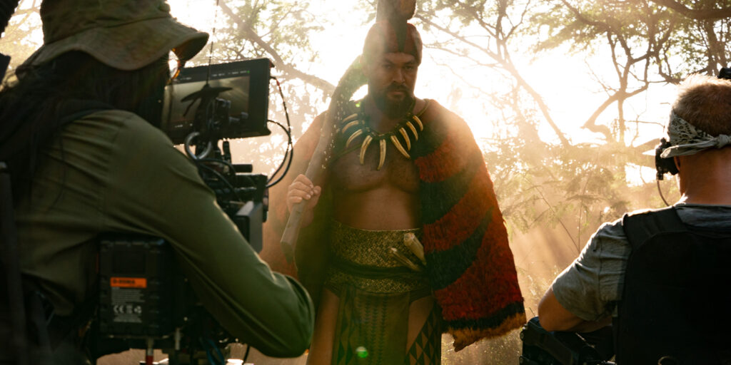 A film crew records an actor in costume, wearing a feathered cloak and large necklace, on a forest set with sunlight streaming through the trees as another long year of production closes. Camera equipment and crew members are visible in the foreground.