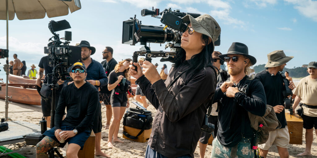 A group of people work with film equipment on a sandy beach as a long year of filming closes. In the foreground, someone operates a camera rig while others stand nearby, some holding kit under the partly cloudy, sunny sky.