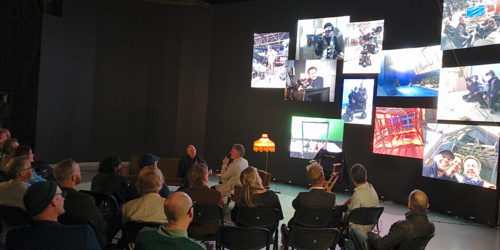A group of people sit facing a dark wall with multiple large screens showing photos of people working on film sets. During this FNF 30th anniversary event, two speakers in front of the audience engage in an insightful discussion.