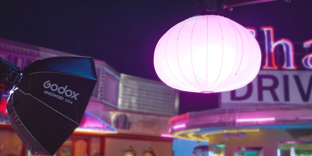 A glowing white paper lantern hangs above a neon-lit street. A high output Godox softbox light is positioned on the left, with blurred signage and colourful lights in the background.