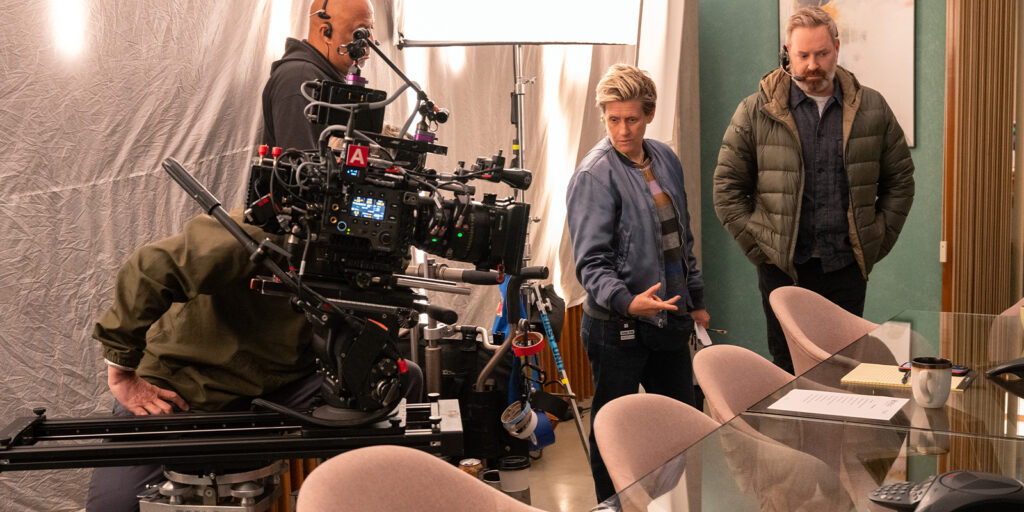 Three people stand on a film set near a large camera and lights, as Jason Oldak gestures towards a conference table with papers, a mug, and other bits and pieces. A fourth person operates the camera.