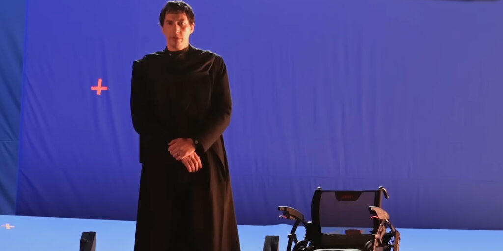 A person in a long, dark robe stands with hands folded in front of a wheelchair on a blue screen set, with two orange tracking markers visible on the background—a scene reminiscent of something from a Mike Figgis megadoc production.