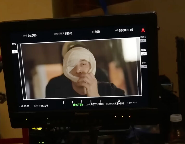 A monitor displays a video recording of a person holding a white cloth or bandage to their face, set against a backdrop of various equipment in a dimly lit room—evoking the documentary style seen in Megadoc by Mike Figgis.
