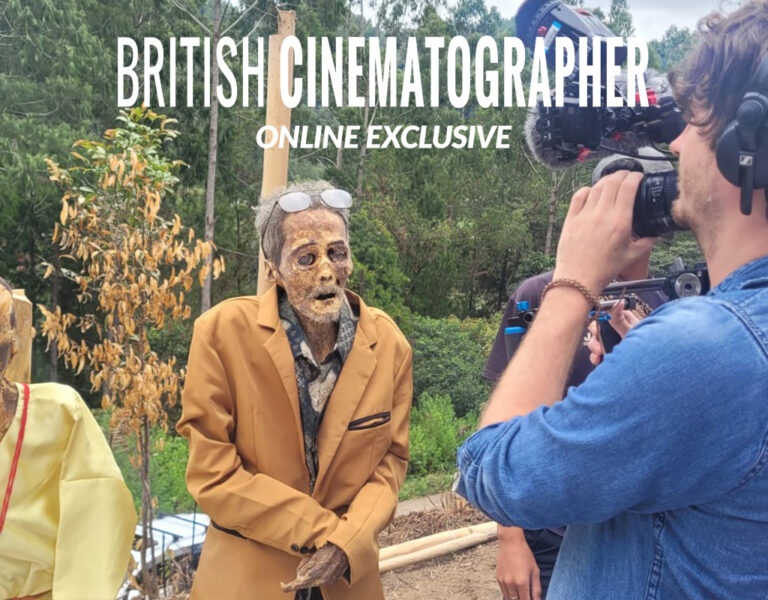 A cameraman films two preserved mummies dressed in clothes and spectacles outdoors, possibly for "Death with Kris," with several people and trees in the background. The text “BRITISH CINEMATOGRAPHER EXCLUSIVE” appears at the top of the image.