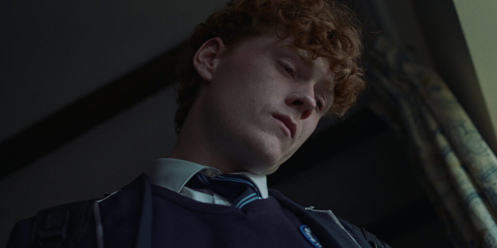 A young person with curly red hair, wearing a school uniform and tie, is seen from a low angle indoors. Their serious expression stands out against patterned curtains—a striking scene inspired by BSC Short Film Competition 2025 winners.