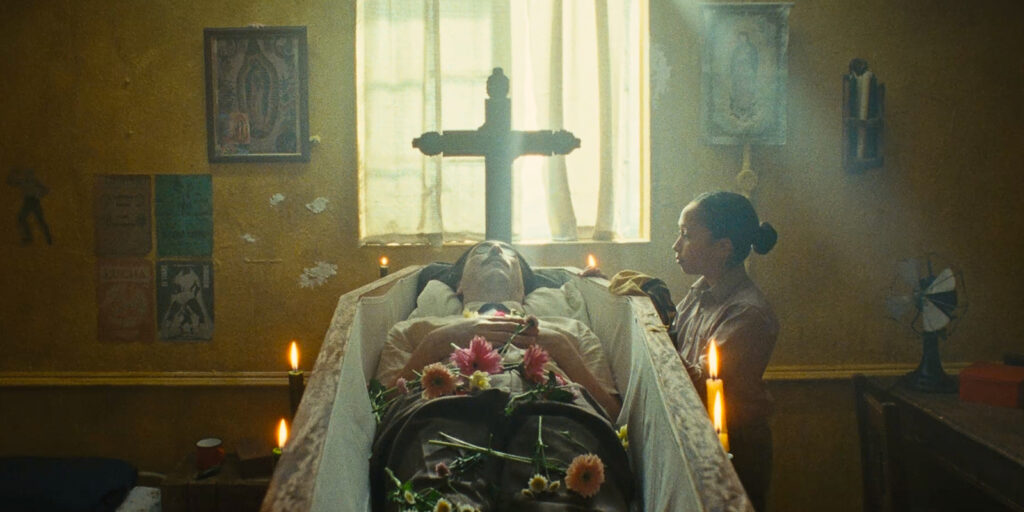 A person lies in a coffin surrounded by flowers and candles, while another stands beside them. Light streams through a curtained window, illuminating a cross and religious images—capturing the poignancy fit for a BSC Short Film Competition entry.