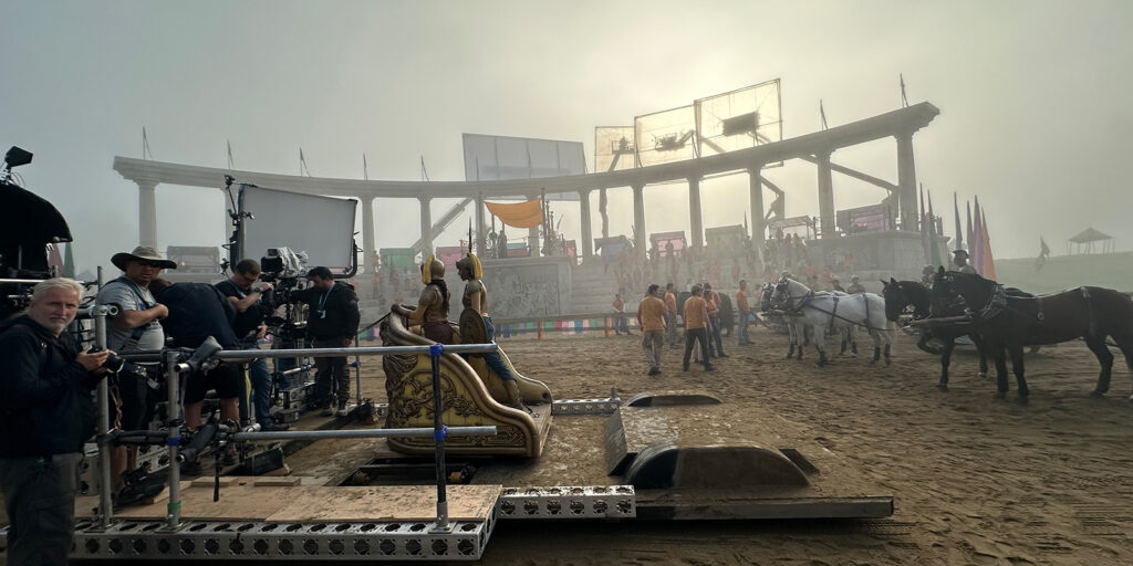 Film crew and camera equipment are set up on a sandy arena with a chariot, horses, and actors in costume. The large Roman-style set evokes an epic scene from Percy Jackson and the Olympians, with atmospheric lighting by Jules O'Loughlin BSC ACS.