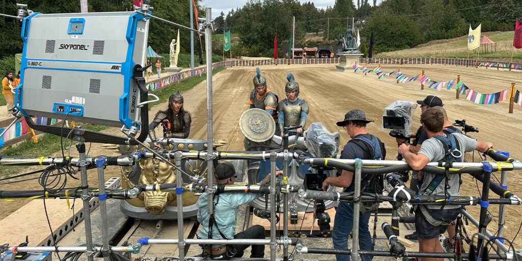 A film crew records actors in Roman-style armour on a set designed to look like an ancient arena, reminiscent of scenes from Percy Jackson and the Olympians, surrounded by production equipment, lights, cameras, colourful flags, and a statue.