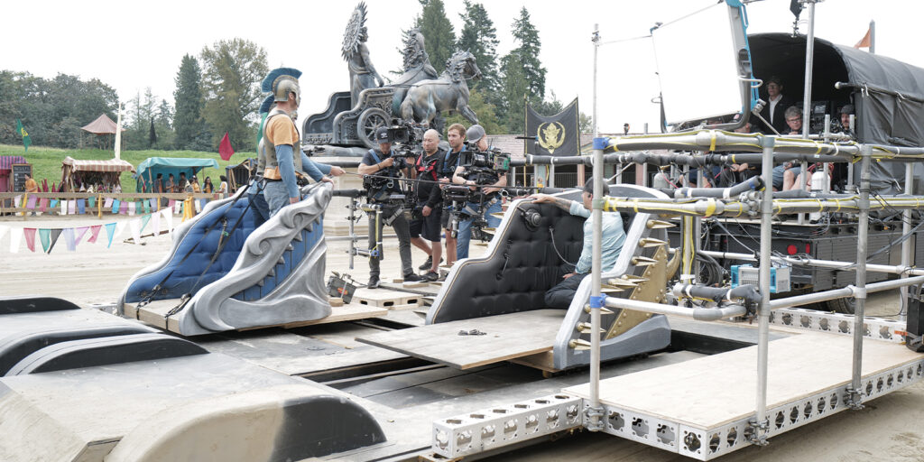 A film crew operates cameras and equipment on a set designed to simulate a chariot scene, with an actor in costume—reminiscent of Percy Jackson and the Olympians—standing on a decorated platform as Jules O'Loughlin directs behind the scenes.