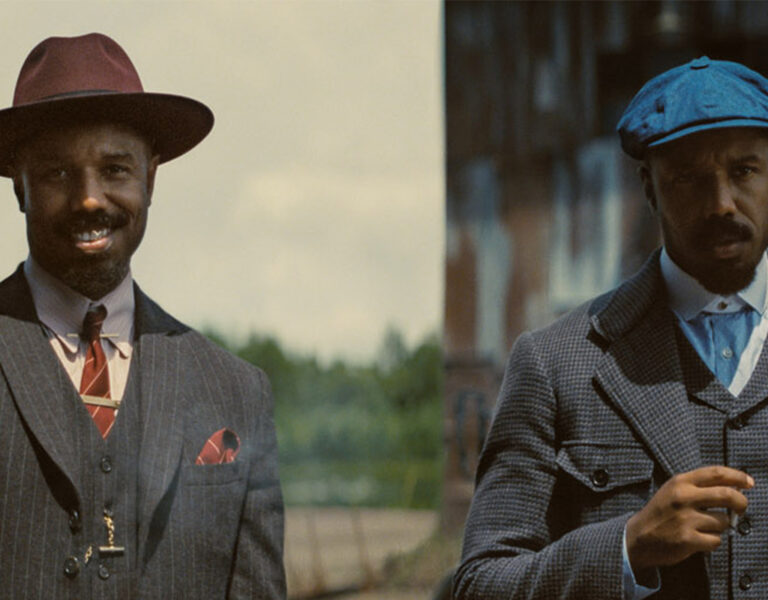 Two men stand side by side, each wearing early 20th-century suits, evoking a scene worthy of the National Board of Review. The man on the left sports a maroon hat and striped suit; the man on the right, a blue cap and checked suit, holds an object in his hand.