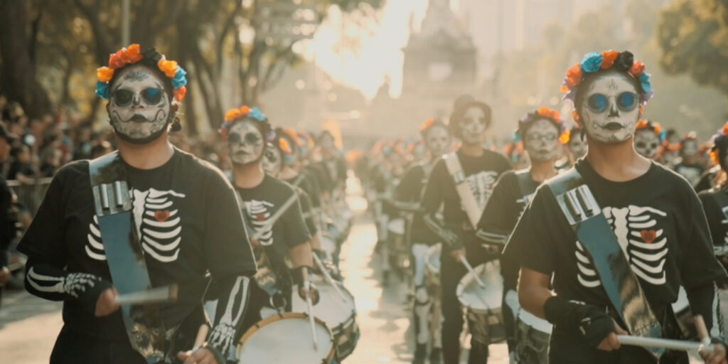 A parade with people wearing skeleton costumes, face paint resembling skulls, and flower crowns—reminiscent of Death with Kris and Andy Pritchard—playing drums whilst walking down a street lined with spectators.