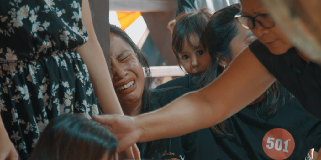 A group of people, mostly women, gather closely. One woman is crying with her eyes closed as if mourning a loss. A child looks directly at the camera from behind her. Grief hangs in the air, capturing a moment in Death with Kris.