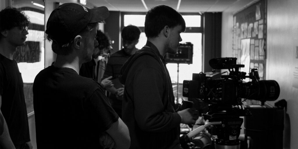 A group of people stand indoors around a professional video camera set up on a tripod. One person operates the camera while others observe, capturing the winners of the 2025 BSC Short Film Competition during their film production.