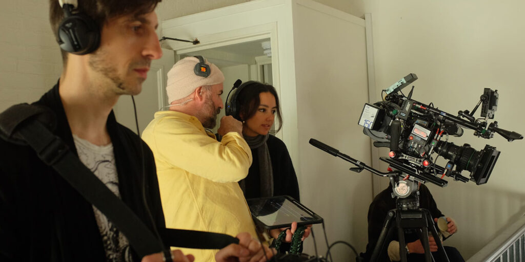 Three crew members work on a film set; one holds audio equipment, another in a yellow shirt gives instructions, and a third person stands near a large professional camera mounted on a tripod.