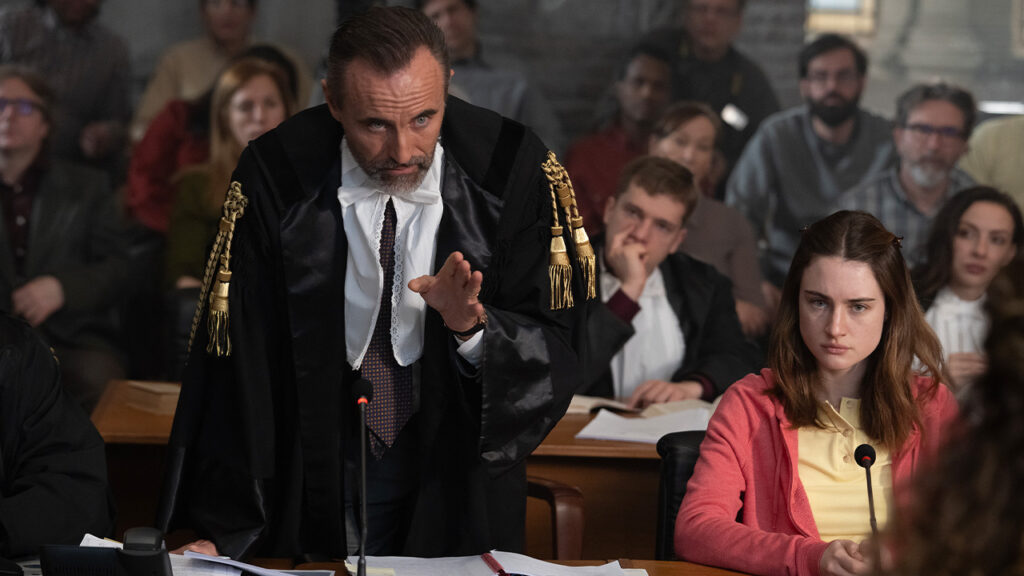 A man in a black robe stands and gestures whilst speaking in a courtroom. Beside him, Bella Gonzales in a pink cardigan sits with a serious expression. In this twisted tale, several people watch attentively from the background.