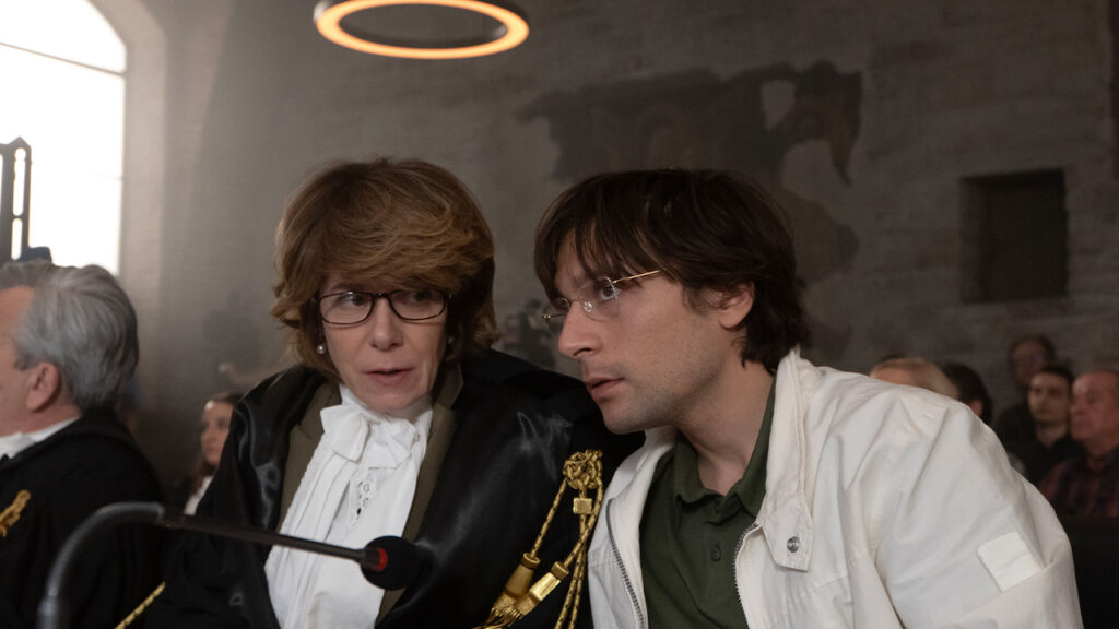 Two people sit close together indoors—one in legal robes and spectacles, the other casually dressed—engaged in a serious conversation. Reminiscent of a scene from a Twisted Tale involving Amanda Knox or Bella Gonzales, others watch in the background.