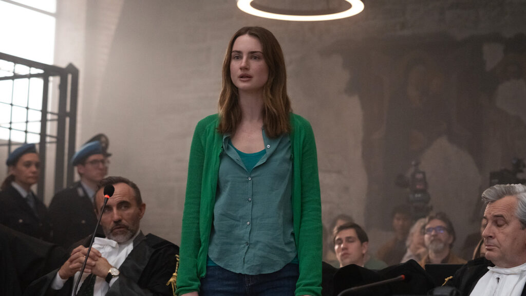 A young woman in a green cardigan, resembling Bella Gonzales, stands in a courtroom surrounded by lawyers and officials, with a circular light above her and a large mural on the wall—a scene reminiscent of The Twisted Tale of Amanda Knox.