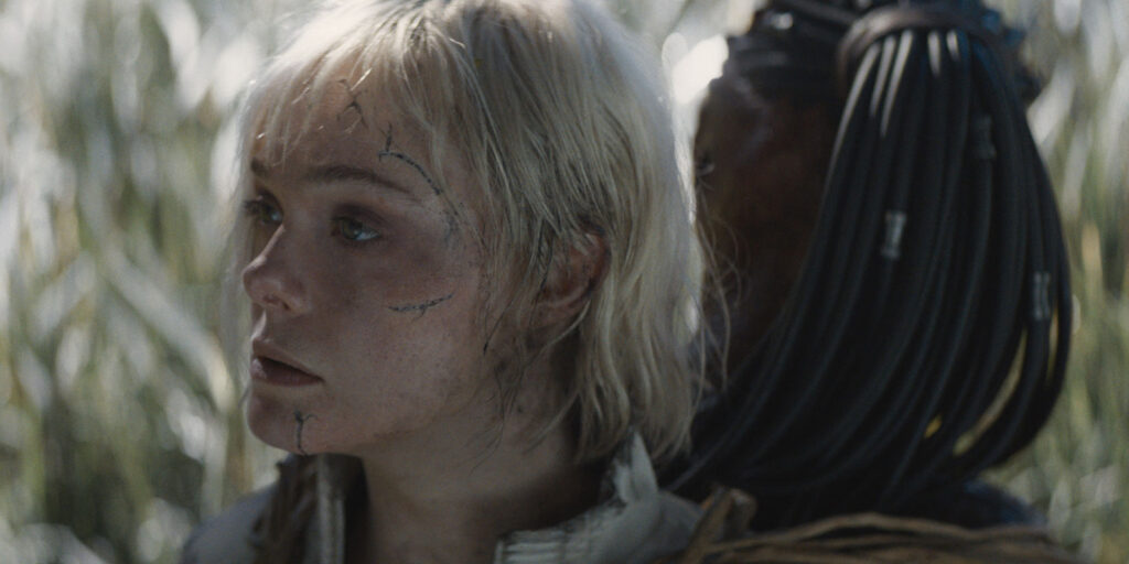 A person with messy blonde hair and scratch marks on their face stands in front of someone in dark armour and cables, evoking scenes from Predator: Badlands. Tall grass and a blurred background complete the tense atmosphere.