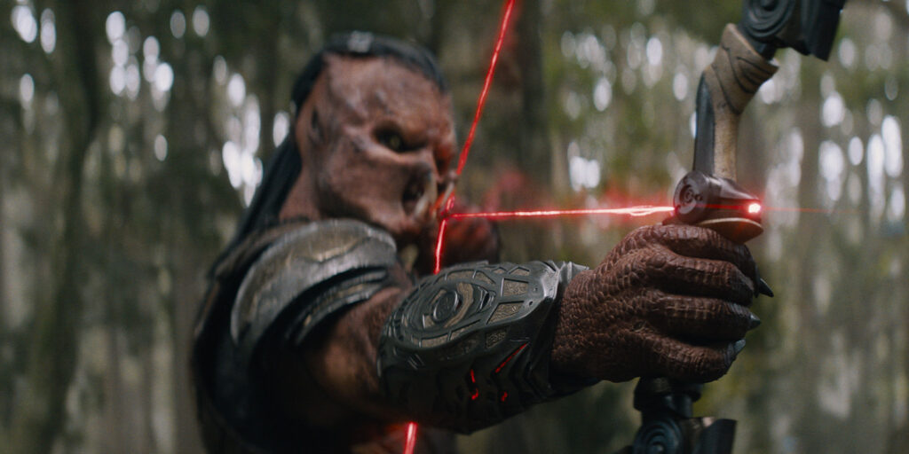 A creature with textured skin and armour draws a bow with three red laser sights in a forest setting. Channelling Jeff Cutter’s cinematic style, this close-up captures the hand, bow, and intense expression reminiscent of Predator: Badlands.