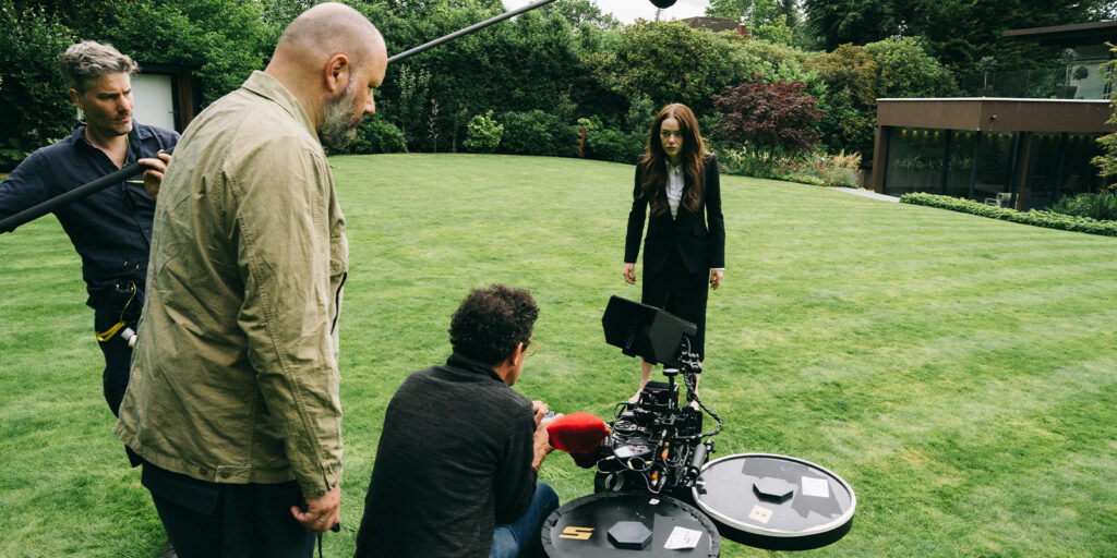 Three crew members operate camera equipment on a grassy lawn, capturing the positives in the negative as a woman in a black suit stands before them. In the background, trees and a modern house complete the scene.