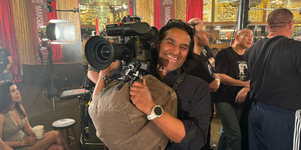 A cameraman smiles whilst holding a professional video camera on his shoulder at an indoor event. Several people are in the background, and glittery decorations and bright lights are visible.