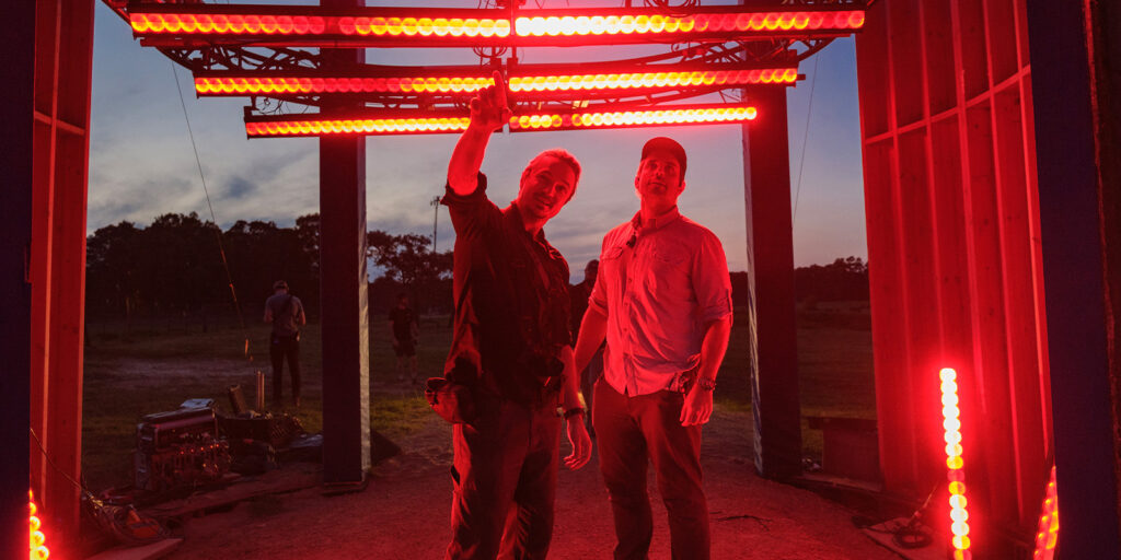 Two people stand under a structure with bright red lights outdoors at dusk, evoking a Stranger Things vibe. One points upwards while the other stands beside him; equipment and trees are visible in the background.