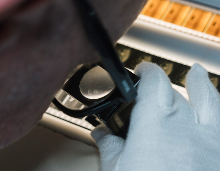 A person wearing white gloves examines photographic film negatives on a light table using a magnifying glass, carefully checking the illuminated strips for preservation. Their face is partially visible as they work to protect these historical images.
