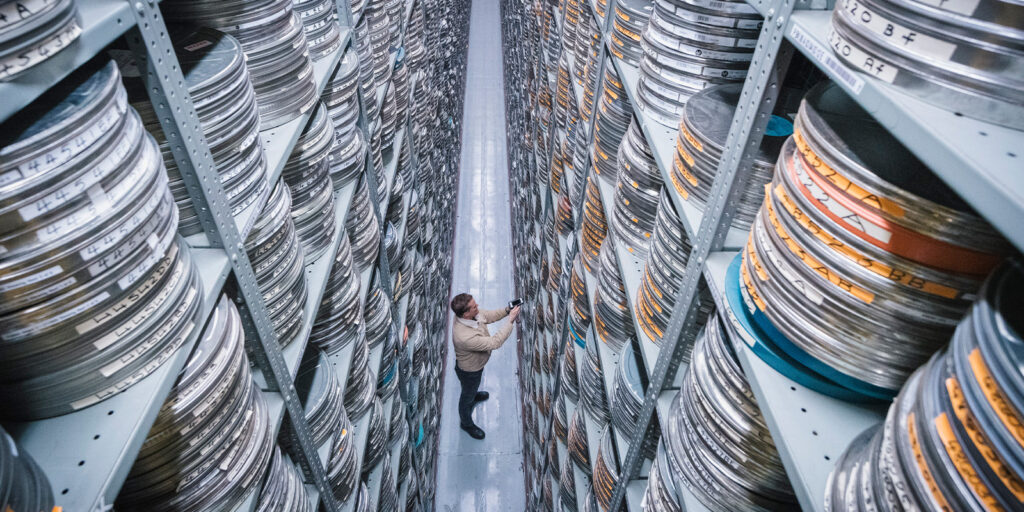 A person stands in a narrow aisle between tall shelves filled with metal film canisters, reaching up to retrieve a reel—part of the ongoing preservation and restoration efforts to protect cinematic history stored on these towering shelves.
