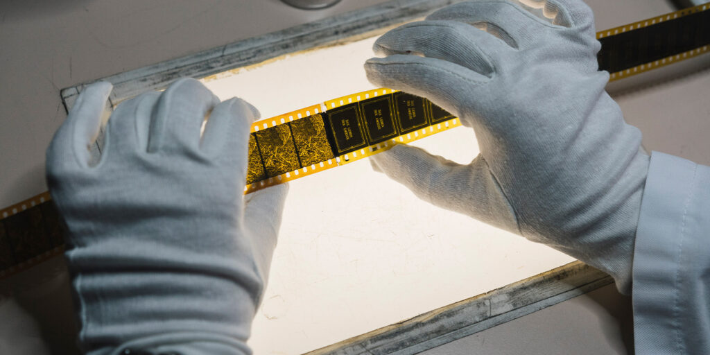 A person wearing white gloves is carefully examining a strip of photographic film negative on a lightbox, emphasising the importance of preservation and restoration.