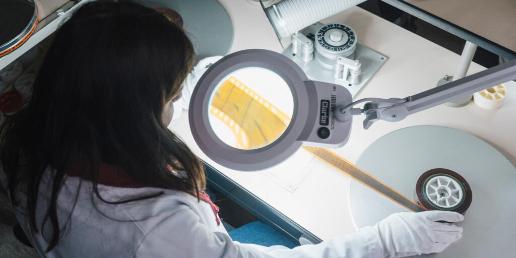 A person in a white lab coat and gloves examines a strip of photographic film under a magnifying lamp, carefully working on film restoration at a worktable filled with reels and equipment.