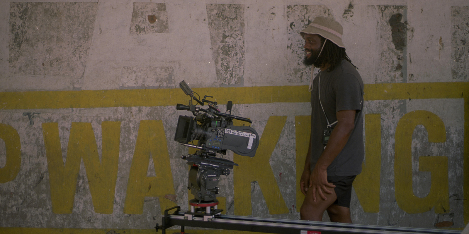 A person stands smiling next to a large professional video camera mounted on a track in front of a weathered wall with faded yellow text. The individual wears a bucket hat, T-shirt, and shorts.