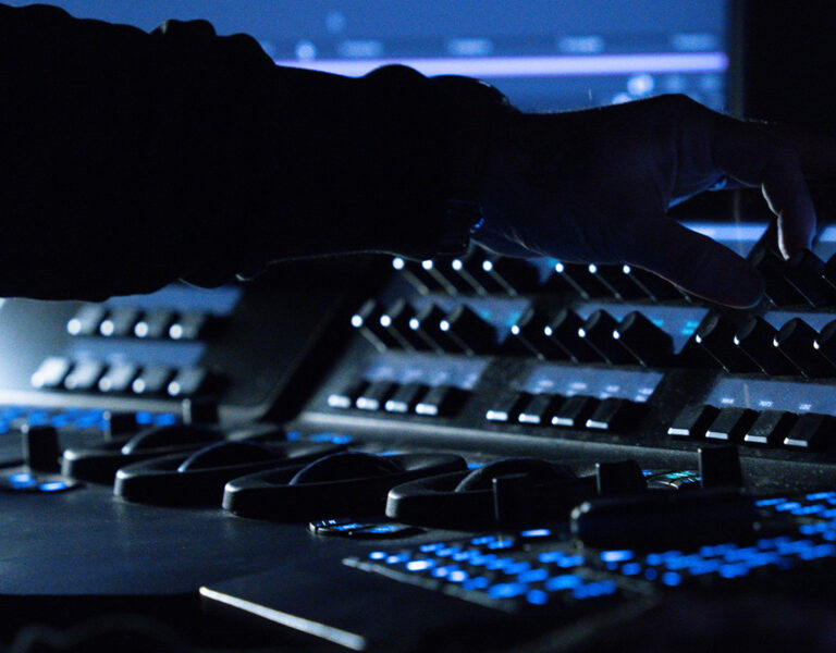 A person adjusts knobs and controls on a professional audio or video editing console in a dimly lit film lab, with illuminated buttons and screens visible—capturing the essence of film development.