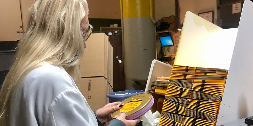 A person with long blonde hair examines a round film canister next to a stack of brown envelopes in an industrial setting, finding positives in the negative amid cardboard boxes and office equipment in the background.