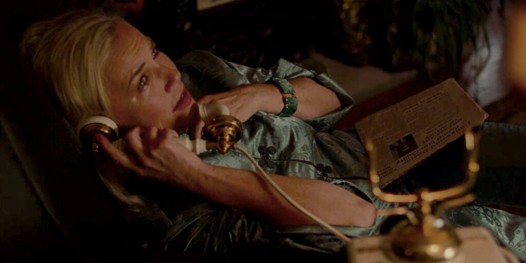 A woman with fair hair in a blue dressing gown is lying down, holding a vintage telephone to her ear and a folder in the other hand, as if engaged in lab collaboration. She appears to be in conversation and is looking upwards.