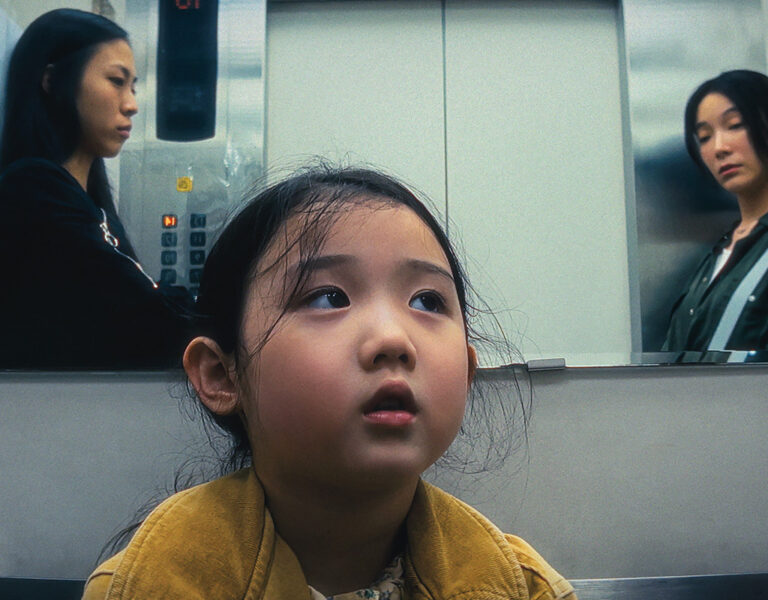A young girl with a yellow jacket looks upward in a lift. Two women, one in black and one in green, are reflected in the mirror behind her. Text above reads, British Cinematographer Online Exclusive: Left-Handed Girl by Tzu-Hao Kao.