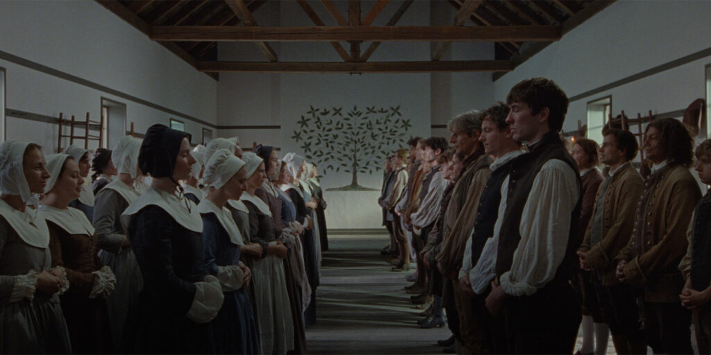 A group of men and women in period clothing stand facing each other in two lines inside a large, wooden-beamed hall with a wall decorated with a white tree and black birds in the background.