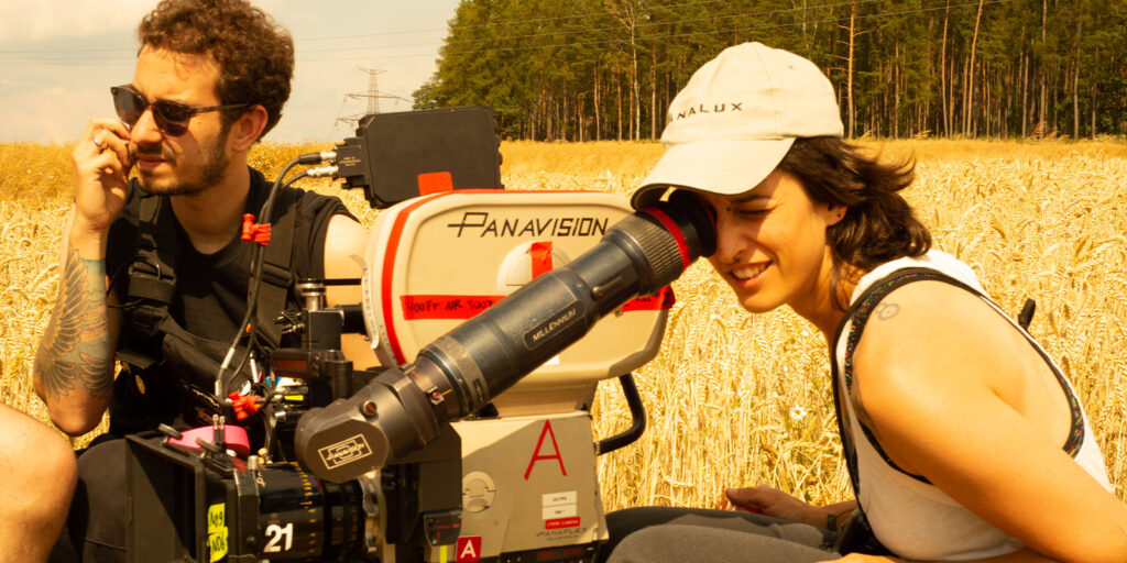Two people operate a large Panavision film camera in a wheat field on a sunny day. One looks through the camera’s viewfinder while the other speaks into a walkie-talkie. Trees and power lines are visible in the background.