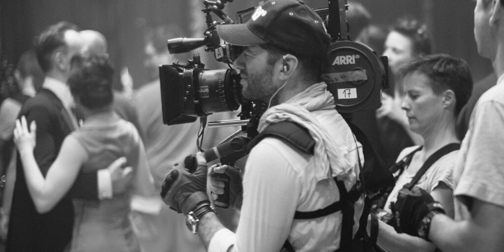 A camera operator wearing a cap and harness films a scene with people dancing in the background. Another crew member stands nearby. The image is in black and white.