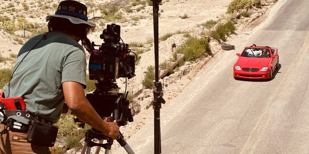 A person operating a camera on a tripod films a red convertible car driving down a deserted, sunlit road with sparse vegetation on the sides.