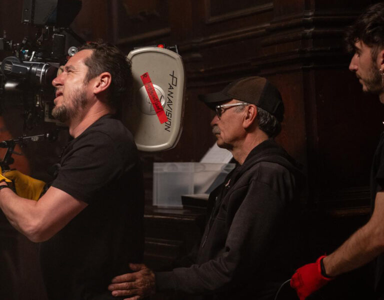 Three crew members on a film set operate a camera; one in yellow gloves, another in a cap and glasses, and a third dressed in black watch closely—all working together amid wood panelling, emphasising sustainability behind the scenes.