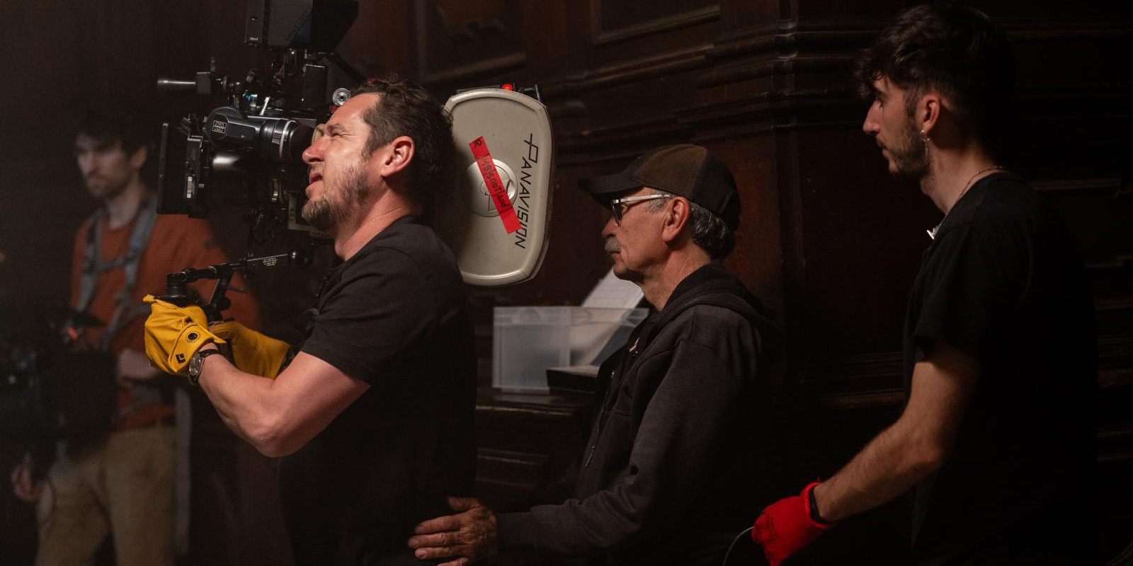 Three crew members on a film set operate a camera; one in yellow gloves, another in a cap and glasses, and a third dressed in black watch closely—all working together amid wood panelling, emphasising sustainability behind the scenes.