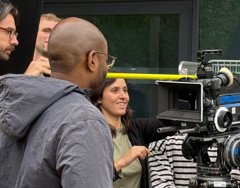 A group of people stand around a professional film camera outdoors. One person adjusts the camera, whilst others observe or discuss, suggesting a collaborative filming or production setting.