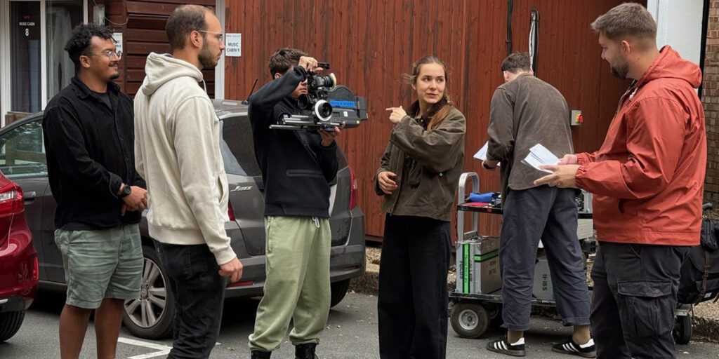 Five people stand outdoors near parked cars and equipment. One person holds a film camera, another gestures while speaking, and the others observe or interact with papers. They appear to be involved in a film production.