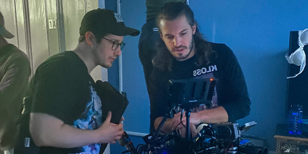 Two men, Ethan Evans and Marti Guiver, stand close together indoors, focused on a monitor atop a professional video camera set-up. One wears glasses and a cap; the other sports long hair and a KLOSS shirt. The room glows with blue-hued light, shutting out outside noise.