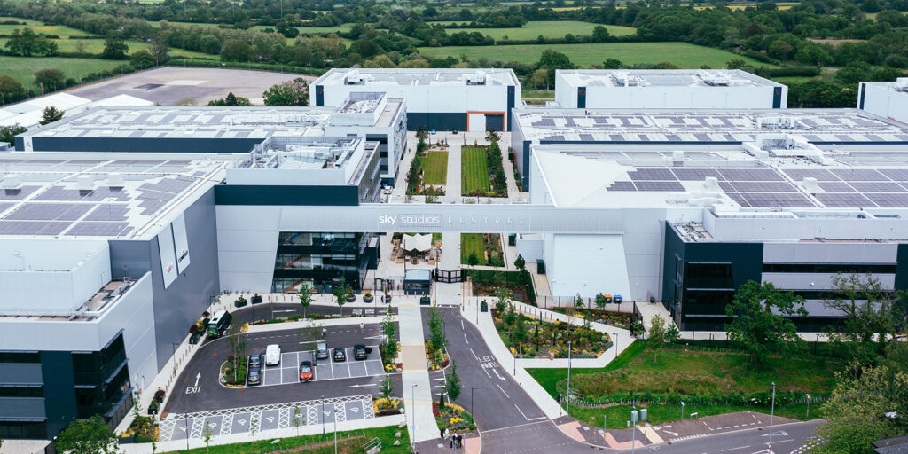 Aerial industry wide shot of the large modern Sky Studios Elstree complex, featuring multiple white buildings with solar panels, a central courtyard, and lush surrounding greenery.