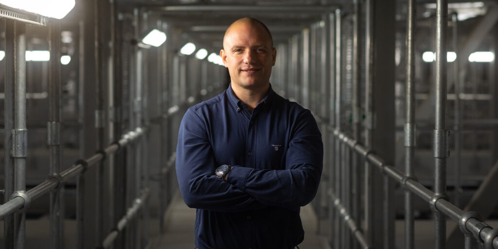 A man with a shaved head wearing a navy blue button-up shirt stands with arms crossed in an industrial setting, captured in an industry wide shot with metal railings and overhead lights, evoking the atmosphere of modern studios.