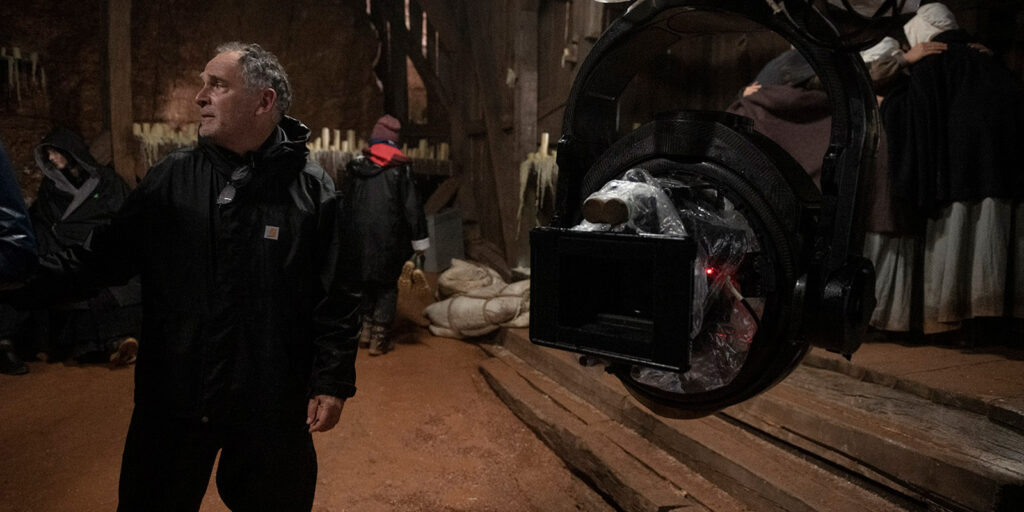 A man in a dark jacket stands in a dimly lit wooden structure with several people in the background. In the foreground, William Rexer ASC's professional film camera—used on The Testament of Ann Lee—is partially covered with polythene.