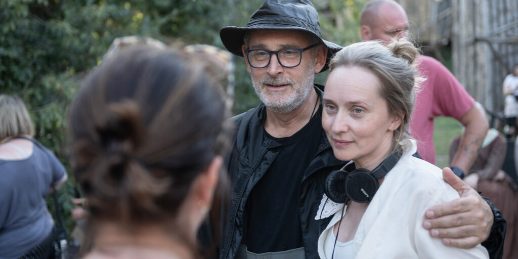 A man wearing glasses and a bucket hat stands with his arm round a woman with headphones round her neck. They are outdoors, with other people and greenery visible, evoking the visual storytelling of William Rexer ASC.